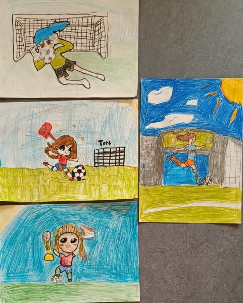 Fünf farbenfrohe Kinderzeichnungen zeigen fußballspielende Mädchen: eine Torhüterin, die taucht, eine Stürmerin, die schießt, eine Spielerin, die einen Ball ins Netz schießt, ein Mädchen, das eine Trophäe und eine Fahne hält, und eine, die einen Ball schießt. Alle sind in sportlichen Outfits auf grünen Feldern mit blauem Himmel zu sehen.