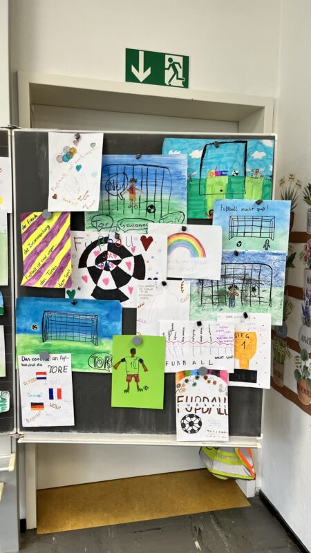 Eine Tafel ist mit farbenfrohen Kinderzeichnungen und -bildern bedeckt, die sich meist um Fußball, Regenbögen und Herzen drehen. Einige zeigen Fußballtore, -felder und -spieler; ein grünes Notausgangsschild befindet sich über der Tafel. Die Umgebung scheint ein Klassenzimmer zu sein.