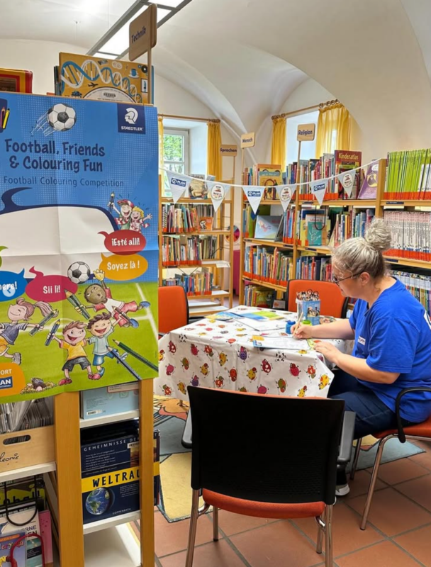 Eine Frau sitzt an einem runden Tisch in einer farbenfrohen Bibliothek und ordnet oder malt Bücher aus. Der Tisch ist mit einem weißen Tuch mit roten Blumen bedeckt. Hinter ihr stehen Regale mit Kinderbüchern. Auf der linken Seite wirbt ein Aufsteller für einen Malwettbewerb zum Thema Fußball.