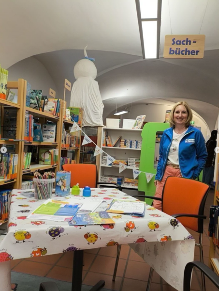 Eine lächelnde Frau steht in einer farbenfrohen Bibliothek neben Bücherregalen und einem Schild mit der Aufschrift "Sachbücher". Auf einem runden Tisch mit Marienkäfer-Muster liegen Papiere, Stifte und Spielzeug. Banner und ein großes weißes Modell hängen in dem hellen, einladenden Raum.