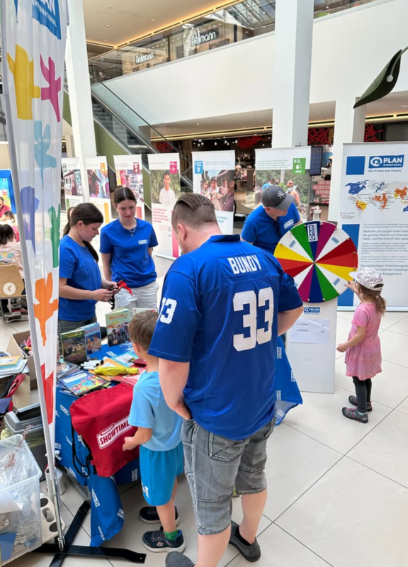 Eine Gruppe von Erwachsenen und Kindern interagiert an einem Veranstaltungsstand mit einem farbenfrohen Preisrad, Postern und Tischen mit Informationsmaterial. Die Mitarbeiter tragen passende blaue Hemden. Ein Mann mit der Aufschrift "BUNDY 33" auf seinem Hemd steht neben zwei kleinen Kindern.