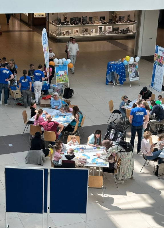 Eine Gruppe von Erwachsenen und Kindern sitzt an Tischen in einem geräumigen Innenbereich und beschäftigt sich mit Bastelarbeiten und Aktivitäten. Freiwillige Helfer in blauen Hemden helfen ihnen dabei. Im Hintergrund sind bunte Luftballons, Plakate und Informationsbanner zu sehen, während die Menschen spazieren gehen und beobachten.