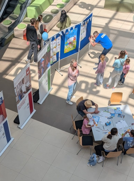 Eine Draufsicht auf eine öffentliche Veranstaltung in einer Halle zeigt Menschen, darunter auch Kinder, die sich an Tischen mit Aktivitäten beschäftigen. Kunstwerke sind auf Tafeln ausgestellt, und in der Nähe stehen Informationsbanner. Einige Leute malen oder zeichnen, während andere beobachten oder herumlaufen.
