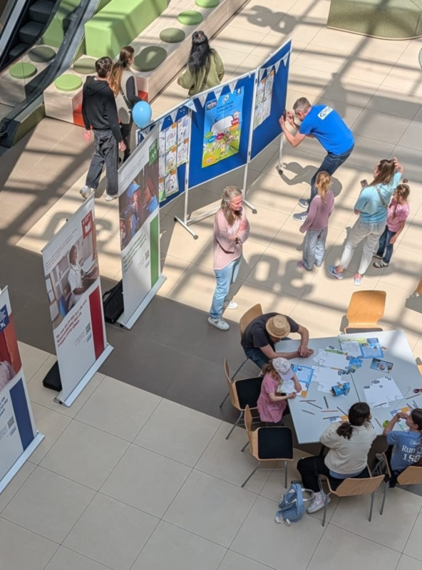 Eine Draufsicht auf eine öffentliche Veranstaltung in einer Halle zeigt Menschen, darunter auch Kinder, die sich an Tischen mit Aktivitäten beschäftigen. Kunstwerke sind auf Tafeln ausgestellt, und in der Nähe stehen Informationsbanner. Einige Leute malen oder zeichnen, während andere beobachten oder herumlaufen.