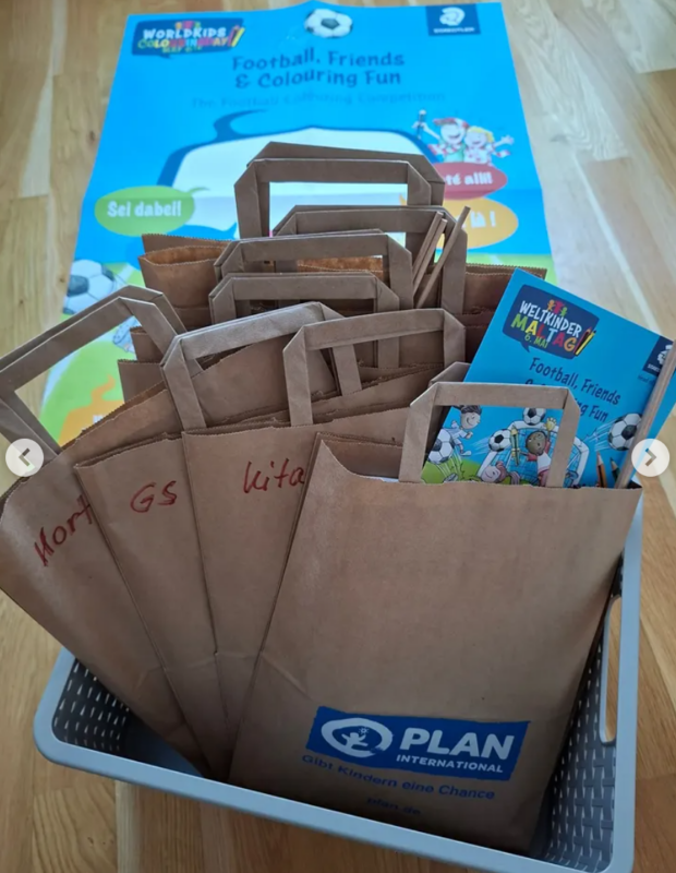 Ein Plastikkorb enthält mehrere braune Papiertüten mit Henkeln und handgeschriebenen Etiketten. Einige Tüten enthalten bunte Hefte mit dem Titel Fußball, Freunde und Malspaß. Im Hintergrund ist ein großes blau-gelbes Poster mit demselben Thema zu sehen.