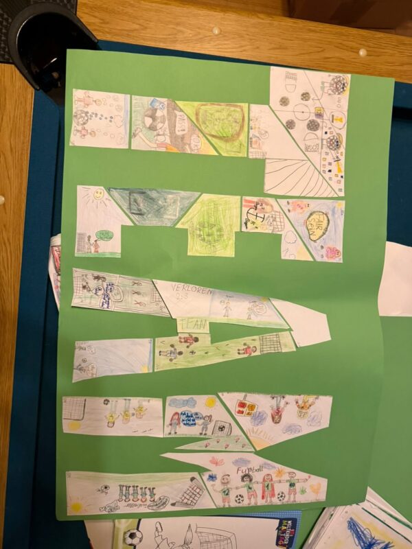 Ein großes grünes Poster zeigt ausgeschnittene Formen, die mit bunten Kinderzeichnungen gefüllt sind, darunter Sportszenen, Menschen, Fußballnetze, Bälle, Gesichter, Tiere und verspielte Kritzeleien. Die Formen sind in einem geometrischen Muster auf einem Holztisch angeordnet.