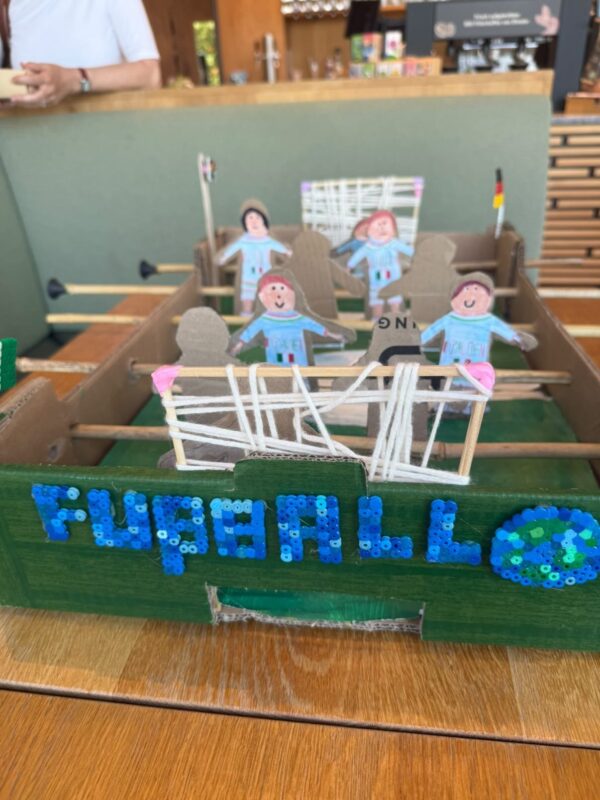 Ein handgefertigtes Tischfußballspiel aus Pappe und Holz mit der Aufschrift FÜR ALLE in blauen Perlen an der Seite. Die Spielerfiguren sind Pappfiguren mit lächelnden Gesichtern, die jeweils ein blaues Trikot tragen. Eine weiße Schnur bildet das Tornetz.