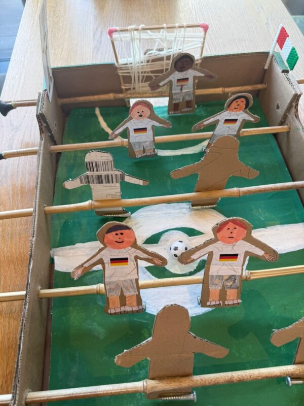 Ein selbstgebautes Tischfußballspiel aus Pappe zeigt handgezeichnete Spieler mit roten Wangen und Deutschlandtrikots. Die Spieler sind an Holzstangen befestigt, und auf der Rückseite ist ein weißes Netztor mit einem kleinen Torpfosten auf einem grün gestrichenen Feld zu sehen.