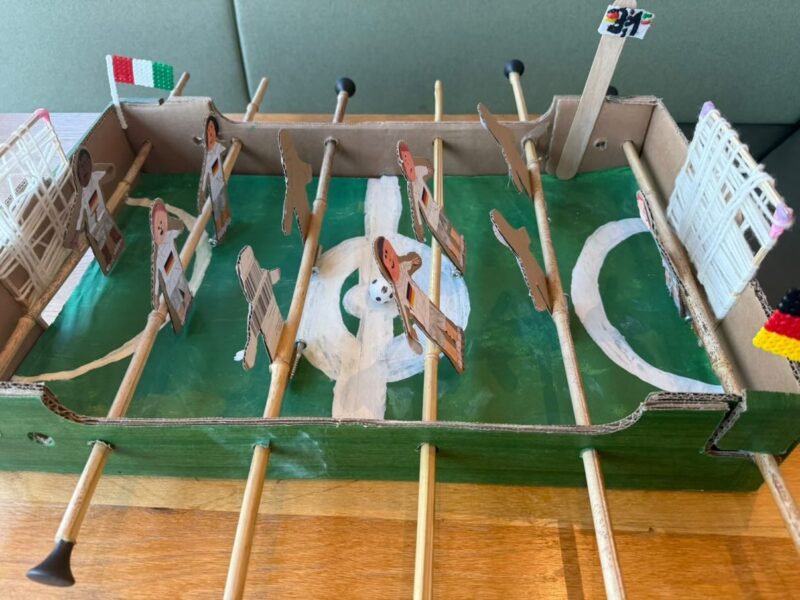 Ein selbstgebauter Tischkicker aus Pappe, Holzdübeln und verzierten Papierfiguren steht auf einer Holzfläche. Das grüne Spielfeld ist mit weißen Markierungen versehen und in den Ecken sind kleine Fähnchen angebracht. In der Mitte des Feldes liegt ein kleiner Fußball.