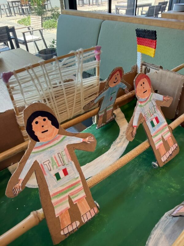 Drei gemalte Pappfiguren von Kindern, die alle Trikots mit der Aufschrift ITALIA und italienischen Flaggen tragen, stehen auf einer grünen Fläche in der Nähe eines behelfsmäßigen Fußballtors aus Pappe und Schnur. Im Hintergrund ist eine kleine deutsche Flagge zu sehen. Das Sonnenlicht fällt durch ein Fenster ein.