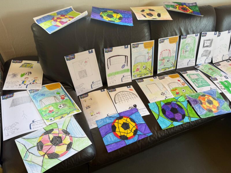 Auf einer schwarzen Couch sind viele bunte Kinderzeichnungen zum Thema Fußball zu sehen, die hauptsächlich Bälle, Tore und Felder zeigen. Die Kunstwerke sind in Reihen auf der Sitzfläche und der Rückenlehne angeordnet, wobei sich einige Blätter überlappen und andere darauf liegen.