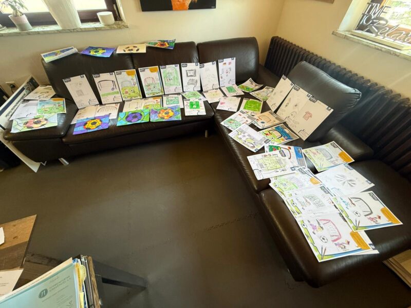 Eine braune L-förmige Couch ist mit vielen bunten Kinderzeichnungen und Arbeitsblättern bedeckt. Die Kunstwerke zeigen geometrische Formen, Häuser, Blumen und kreative Designs. Durch zwei Fenster hinter der Couch strömt Sonnenlicht herein und erhellt den Raum.