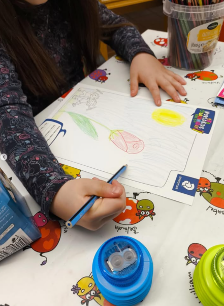 Ein Kind mit langen braunen Haaren malt mit Buntstiften eine Blume, eine Sonne und Gras auf weißem Papier. Auf der Tischdecke sind bunte Cartoon-Insekten abgebildet, und in der Nähe liegen Buntstifte und ein blauer Bleistiftspitzer.