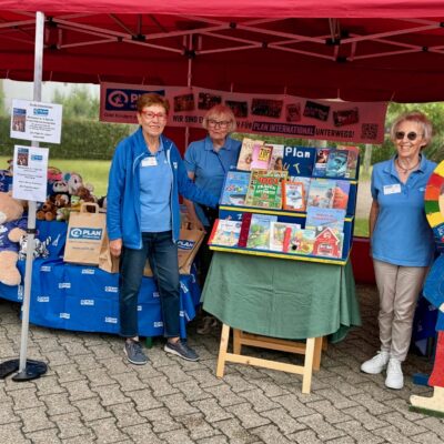 Drei Frauen stehen in einem roten, überdachten Stand mit Bannern von Plan International. Auf Tischen liegen Plüschtiere und Broschüren. Die Frauen tragen blaue Hemden und Namensschilder, und eine steht neben einer bunten Holzfigur eines lächelnden Kindes, das ein Plan-Logo hält.