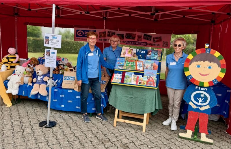 Drei Frauen stehen in einem roten, überdachten Stand mit Bannern von Plan International. Auf Tischen liegen Plüschtiere und Broschüren. Die Frauen tragen blaue Hemden und Namensschilder, und eine steht neben einer bunten Holzfigur eines lächelnden Kindes, das ein Plan-Logo hält.