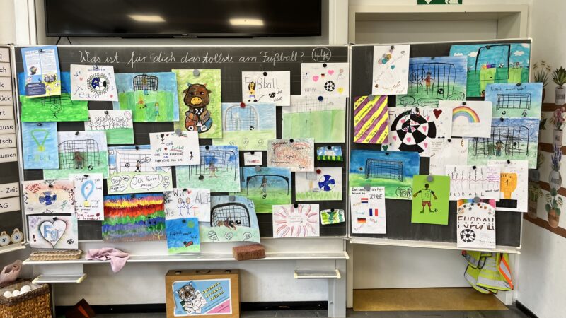 Eine Tafel im Klassenzimmer mit bunten Kinderzeichnungen zum Thema Fußball, darunter Tore, Bälle, Fahnen und Maskottchen. Ein deutscher Text auf der Tafel fragt, was die Schüler am Fußball lieben. Das Klassenzimmer ist hell und aufgeräumt, die Materialien sind unter der Tafel sichtbar.