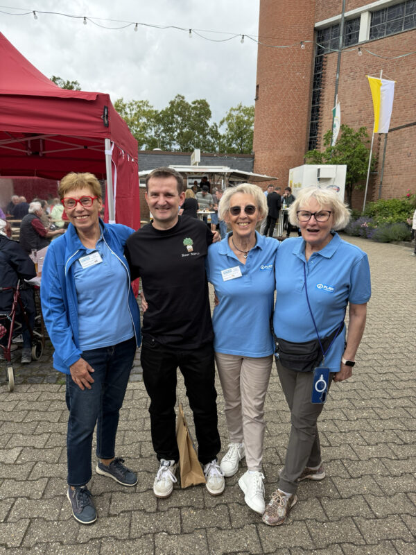 Vier Personen posieren lächelnd im Freien; drei ältere Frauen in blauen Oxfam-Shirts und ein jüngerer Mann in einem schwarzen T-Shirt stehen Arm in Arm vor einem roten Zelt und einem Backsteingebäude. Im Hintergrund sind festliche Dekorationen und Menschen zu sehen.