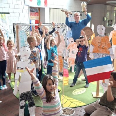 Eine Gruppe von Kindern und ein Erwachsener posieren begeistert in einem Raum, die Hände erhoben, inmitten lebensgroßer, handgezeichneter Papierschnitte von Fußballspielern. Der Erwachsene hält einen Pokal in der Hand. Ein Kind ganz vorne hält eine blau-weiß-rot gestreifte Fahne. Der Raum ist hell und verspielt dekoriert.