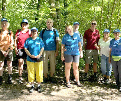 Eine Gruppe von elf Erwachsenen steht draußen auf einem sonnendurchfluteten Waldweg. Die meisten tragen blaue Hemden, Shorts, Wanderstiefel und blaue Mützen. Eine Person trägt eine gelbe Hose. Bäume und Grün umgeben die Gruppe, und Sonnenlicht fällt durch die Blätter über ihnen.