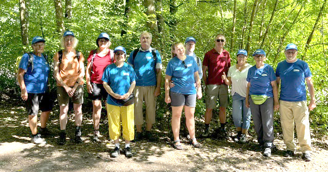 Eine Gruppe von elf Erwachsenen steht draußen auf einem sonnendurchfluteten Waldweg. Die meisten tragen blaue Hemden, Shorts, Wanderstiefel und blaue Mützen. Eine Person trägt eine gelbe Hose. Bäume und Grün umgeben die Gruppe, und Sonnenlicht fällt durch die Blätter über ihnen.