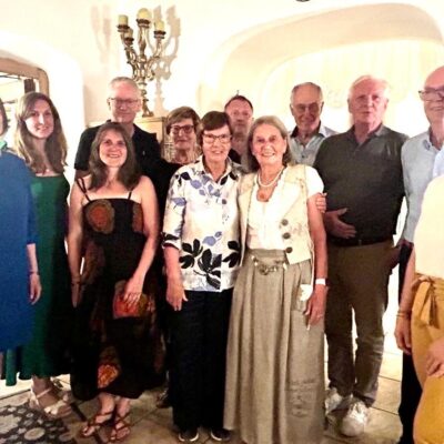 Eine Gruppe von dreizehn Erwachsenen lächelt und posiert gemeinsam in einem Innenraum unter warmer Beleuchtung. Die meisten stehen dicht beieinander; einige haben ihre Arme umeinander gelegt. Der Raum verfügt über gewölbte Türöffnungen, helle Wände und einen Vintage-Kronleuchter, was eine gemütliche, einladende Atmosphäre schafft.