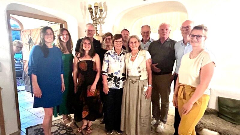 Eine Gruppe von dreizehn Erwachsenen lächelt und posiert gemeinsam in einem Innenraum unter warmer Beleuchtung. Die meisten stehen dicht beieinander; einige haben ihre Arme umeinander gelegt. Der Raum verfügt über gewölbte Türöffnungen, helle Wände und einen Vintage-Kronleuchter, was eine gemütliche, einladende Atmosphäre schafft.