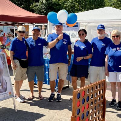Sieben Erwachsene stehen bei einer Veranstaltung im Freien vor einem weißen Zelt. Alle tragen blaue Hemden und lächeln in die Kamera. Zu sehen sind Luftballons, ein „Vier gewinnt“-Spiel und ein Schild mit dem Gesicht eines Kindes und einem deutschen Text. Auf den Tischen liegen Broschüren, Getränke und andere Gegenstände.