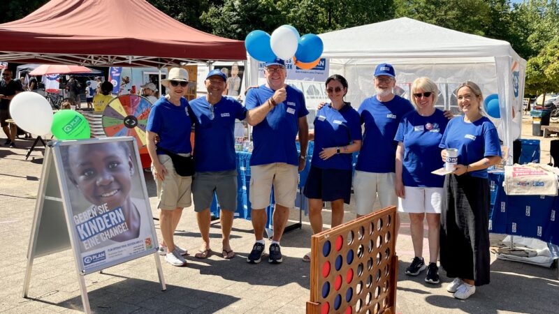 Sieben Erwachsene stehen bei einer Veranstaltung im Freien vor einem weißen Zelt. Alle tragen blaue Hemden und lächeln in die Kamera. Zu sehen sind Luftballons, ein „Vier gewinnt“-Spiel und ein Schild mit dem Gesicht eines Kindes und einem deutschen Text. Auf den Tischen liegen Broschüren, Getränke und andere Gegenstände.