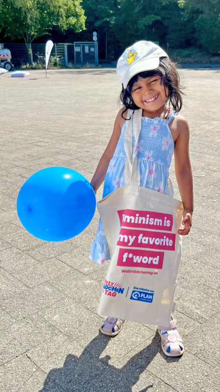 Ein lächelndes junges Mädchen in einem blauen Sommerkleid und einer weißen Mütze steht im Freien und hält einen blauen Luftballon sowie eine beige Tragetasche mit der Aufschrift „Feminism is my favorite F* word“ in der Hand. Im sonnendurchfluteten Hintergrund sind Bäume und ein Zaun zu sehen.
