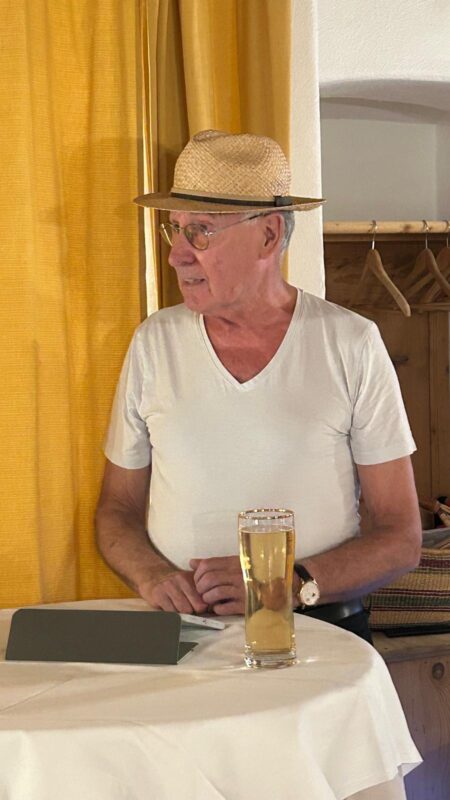 Ein älterer Mann mit Strohhut und weißem T-Shirt steht an einem runden Tisch und hält ein hohes Glas helles Bier in der Hand. Er trägt eine Brille und eine Armbanduhr und steht in der Nähe von gelben Vorhängen und hölzernen Kleiderhaken, wobei er mit entspanntem Gesichtsausdruck nach links blickt.