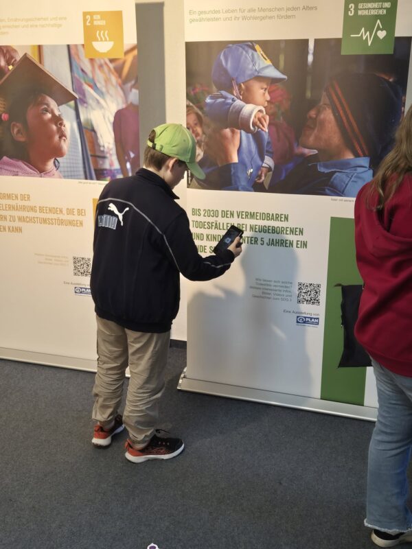 Ein Junge in einer schwarzen Puma-Jacke und einer grünen Mütze scannt einen QR-Code auf einer Ausstellungstafel. Die Ausstellung zeigt Bilder von Kindern und Texte darüber, wie vermeidbare Todesfälle bei Kindern bis 2030 verhindert werden sollen. Eine weitere Person in einem roten Sweatshirt steht in der Nähe, teilweise außerhalb des Bildausschnitts.