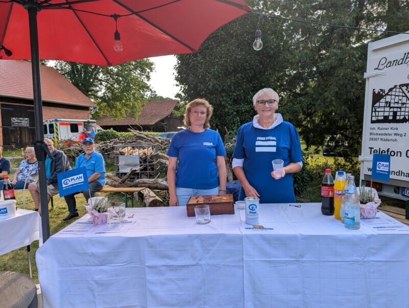 Bei einer Veranstaltung im Freien stehen zwei Frauen in blauen Hemden hinter einem weißen Tisch, auf dem Getränke, eine Spendendose und Flyer liegen, unter einem roten Sonnenschirm. Im Hintergrund sind Bäume, eine Scheune und sitzende Menschen zu sehen. Rechts befindet sich ein weißes Schild mit deutschem Text.