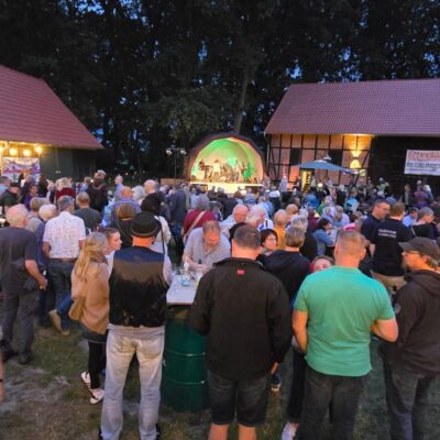 Bei Einbruch der Dämmerung versammelt sich draußen zwischen zwei Scheunen eine große Menschenmenge, die einer Live-Band auf einer kleinen, grün beleuchteten Bühne zusieht. Lichterketten schmücken den Bereich, und Menschen jeden Alters unterhalten sich, stehen beieinander und genießen die Veranstaltung.