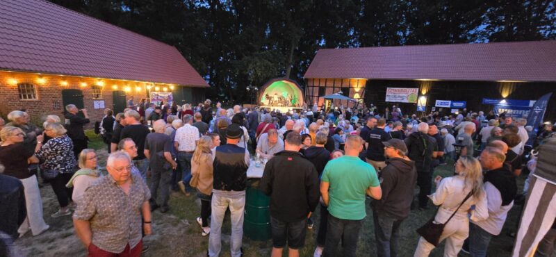 Bei Einbruch der Dämmerung versammelt sich draußen zwischen zwei Scheunen eine große Menschenmenge, die einer Live-Band auf einer kleinen, grün beleuchteten Bühne zusieht. Lichterketten schmücken den Bereich, und Menschen jeden Alters unterhalten sich, stehen beieinander und genießen die Veranstaltung.