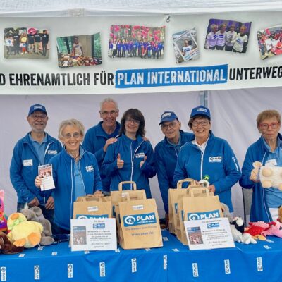 Sieben lächelnde Menschen in blauen Jacken stehen an einem Tisch mit Plüschtieren und Papiertüten unter einem Banner mit der Aufschrift „Wir sind ehrenamtlich für Plan International unterwegs!“. Das Banner ist mit Fotos und einem QR-Code verziert; die Gruppe scheint sich auf einer Wohltätigkeitsveranstaltung zu befinden.