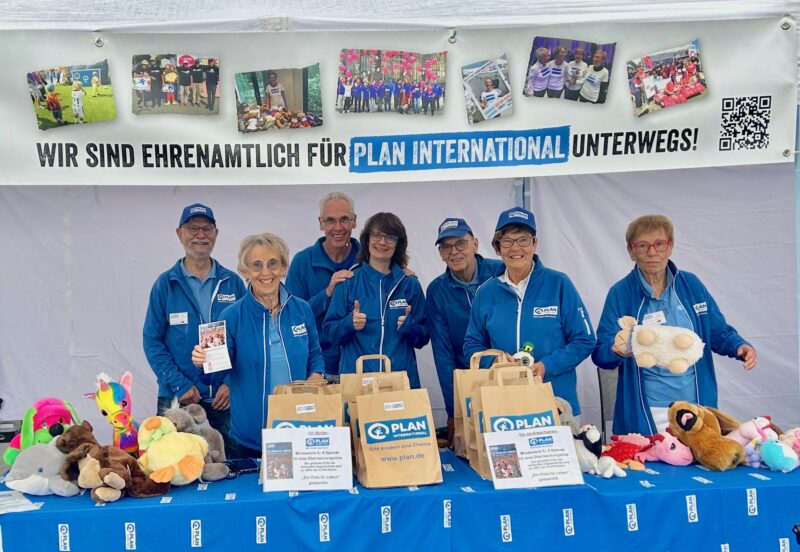 Sieben lächelnde Menschen in blauen Jacken stehen an einem Tisch mit Plüschtieren und Papiertüten unter einem Banner mit der Aufschrift „Wir sind ehrenamtlich für Plan International unterwegs!“. Das Banner ist mit Fotos und einem QR-Code verziert; die Gruppe scheint sich auf einer Wohltätigkeitsveranstaltung zu befinden.
