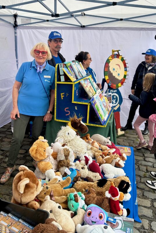 Drei Erwachsene in blauen Uniformen stehen an einem farbenfrohen Spinnrad und einem Bücherregal an einem Stand im Freien. Auf einem Tisch im Vordergrund liegen Plüschtiere. Ein Kind und weitere Personen befinden sich in der Nähe. Die Szene wirkt festlich, unter den Füßen liegt Kopfsteinpflaster.