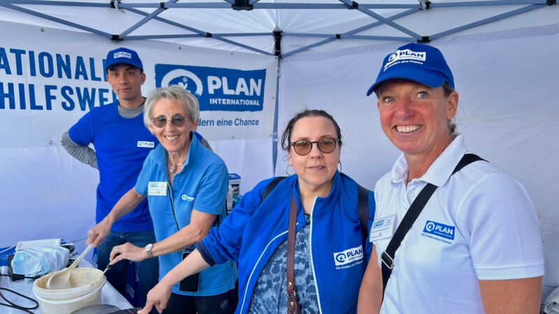 Vier Erwachsene stehen in einer Kabine unter einem Zelt und lächeln in die Kamera. Alle tragen blaue T-Shirts oder Jacken von PLAN International, einige haben auch Mützen auf. Auf einem Tisch vor ihnen liegen Kochutensilien und Lebensmittel. Hinter ihnen hängt ein Banner mit dem Logo von PLAN International.