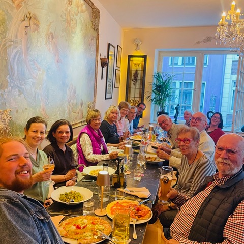 Vierzehn Menschen unterschiedlichen Alters sitzen lächelnd an einem langen Esstisch, der mit Pizza, Pasta und Getränken gedeckt ist, in einem warm beleuchteten Restaurant. An der linken Wand hängen kunstvolle Gemälde, und große Fenster im hinteren Bereich lassen Tageslicht herein. Die Atmosphäre wirkt fröhlich und lebhaft.