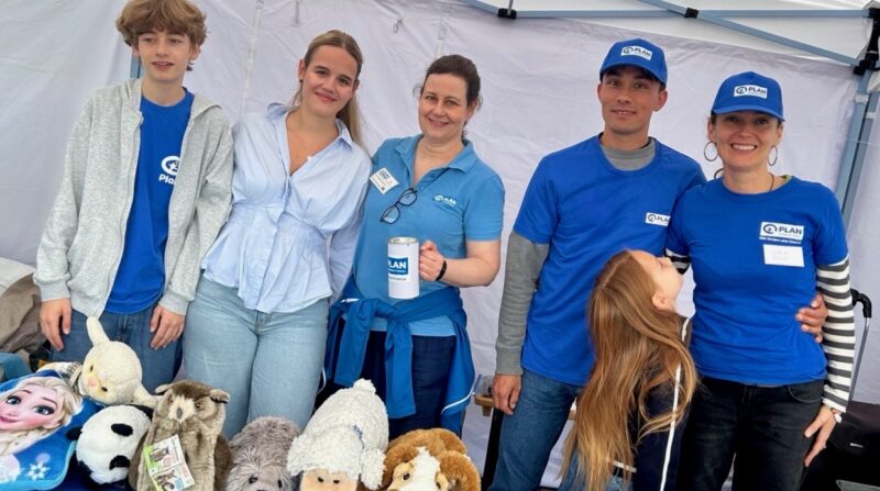 Fünf Erwachsene und ein Kind stehen an einem Stand mit Plüschtieren und einer Elsa-Puppe aus „Die Eiskönigin“. Die meisten tragen blaue PLAN-T-Shirts und Namensschilder. Eine Frau hält einen Becher in der Hand. Der Stand befindet sich unter einem weißen Zelt, und alle lächeln oder unterhalten sich fröhlich miteinander.