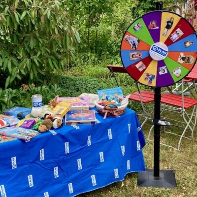 Neben einem Tisch, der mit einem blauen Tischtuch mit dem PLAN-Logo bedeckt ist und auf dem Spielzeug, Notizbücher und Preise ausgestellt sind, steht ein buntes Glücksrad mit Tierfotos. Die Szene spielt sich im Freien ab, umgeben von Grünpflanzen und roten Gartenstühlen.