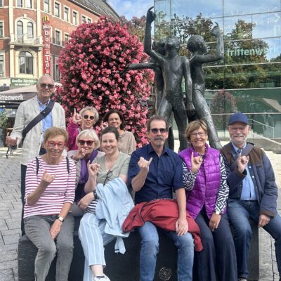 Eine Gruppe von zehn lächelnden Menschen, überwiegend ältere Erwachsene, sitzt und steht vor einer Bronzestatue, die zwei Figuren darstellt. Hinter ihnen befinden sich ein großer rosa blühender Baum, Geschäfte und ein modernes Glasgebäude. Die meisten von ihnen gestikulieren mit erhobenem kleinen Finger und Zeigefinger. Der Tag ist hell und klar.