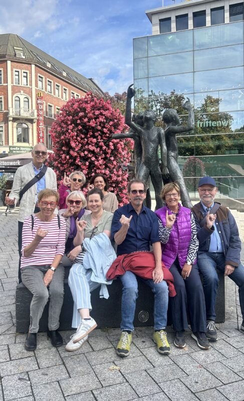 Eine Gruppe von zehn lächelnden Menschen, überwiegend ältere Erwachsene, sitzt und steht vor einer Bronzestatue, die zwei Figuren darstellt. Hinter ihnen befinden sich ein großer rosa blühender Baum, Geschäfte und ein modernes Glasgebäude. Die meisten von ihnen gestikulieren mit erhobenem kleinen Finger und Zeigefinger. Der Tag ist hell und klar.