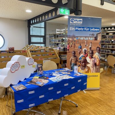 In einer Bibliothek ist ein Ausstellungsstand mit Broschüren, Stiften und Flyern aufgebaut. Dahinter befindet sich ein Standbanner, auf dem lächelnde Kinder und ein Text auf Deutsch zu sehen sind. Im Hintergrund sind Bücherregale und Leseecken zu erkennen. Der Boden ist aus Holz, und die Atmosphäre ist hell und freundlich.