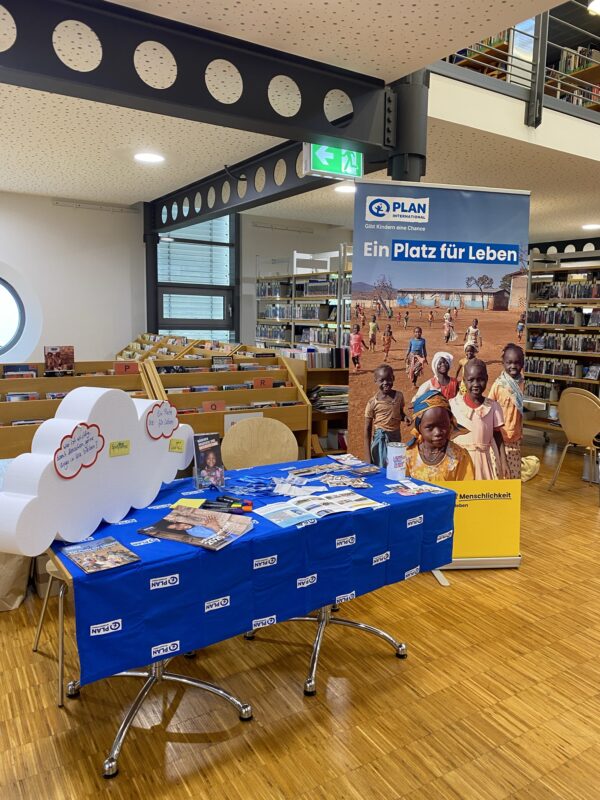 In einer Bibliothek ist ein Ausstellungsstand mit Broschüren, Stiften und Flyern aufgebaut. Dahinter befindet sich ein Standbanner, auf dem lächelnde Kinder und ein Text auf Deutsch zu sehen sind. Im Hintergrund sind Bücherregale und Leseecken zu erkennen. Der Boden ist aus Holz, und die Atmosphäre ist hell und freundlich.