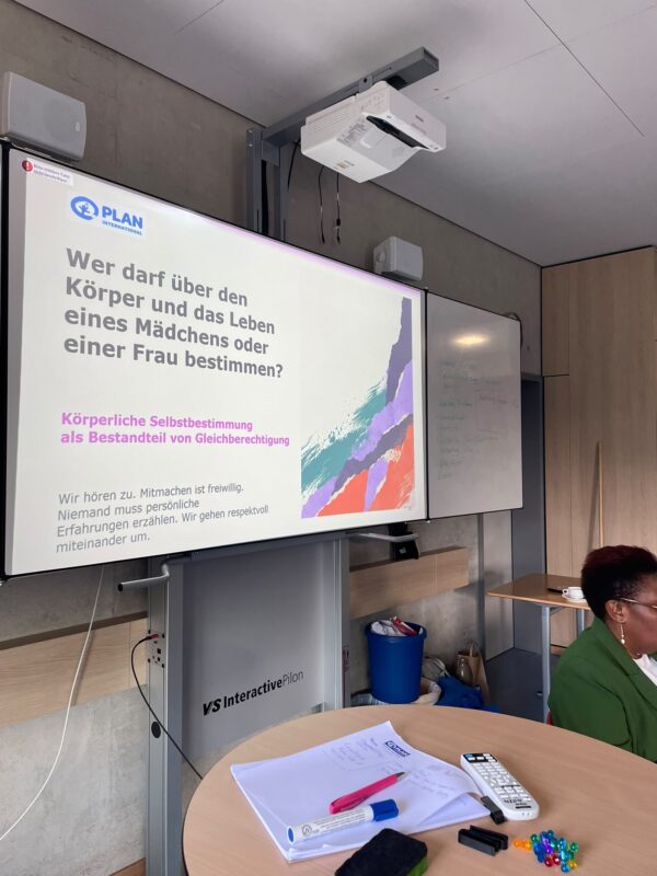 Auf einem großen digitalen Bildschirm ist unter dem Logo von Plan International ein Text in deutscher Sprache zum Thema körperliche Selbstbestimmung von Frauen und Mädchen zu sehen. Im Vordergrund liegen auf einem runden Tisch Papiere und eine Fernbedienung, während rechts davon eine Frau in einem grünen Blazer sitzt.