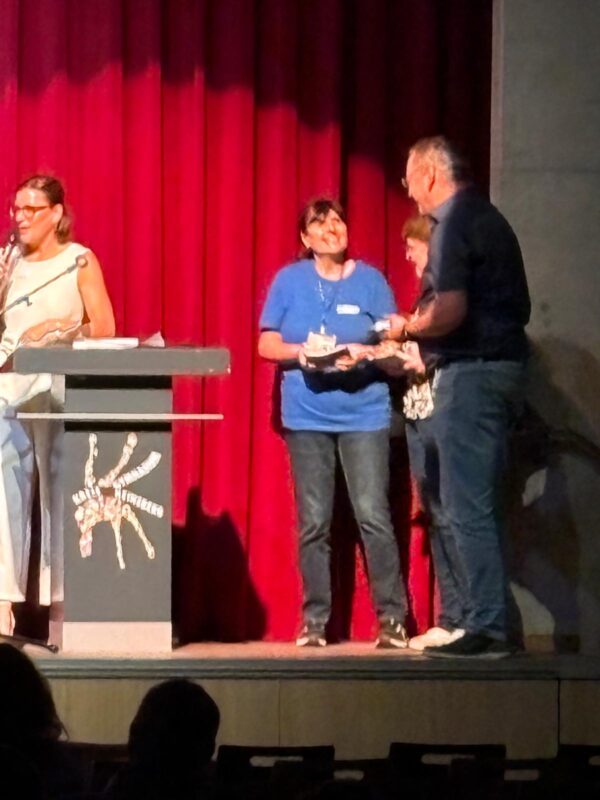 Eine Frau in einem blauen T-Shirt und Jeans steht auf einer Bühne vor roten Vorhängen und lächelt, während sie von einem Mann eine Auszeichnung entgegennimmt. Links steht eine Frau hinter einem Rednerpult, und im dunklen Vordergrund ist ein Teil des Publikums zu sehen.