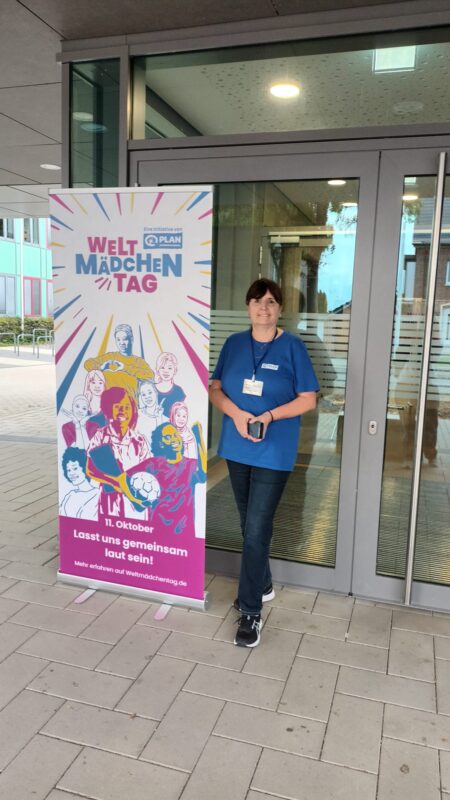 Eine Frau mit dunklen Haaren steht neben einem Werbebanner zum Welt-Mädchentag, auf dem bunte Illustrationen von Mädchen zu sehen sind. Sie trägt ein blaues T-Shirt mit einem kleinen Logo, eine dunkle Jeans und Turnschuhe und posiert vor dem Eingang eines Glasgebäudes.