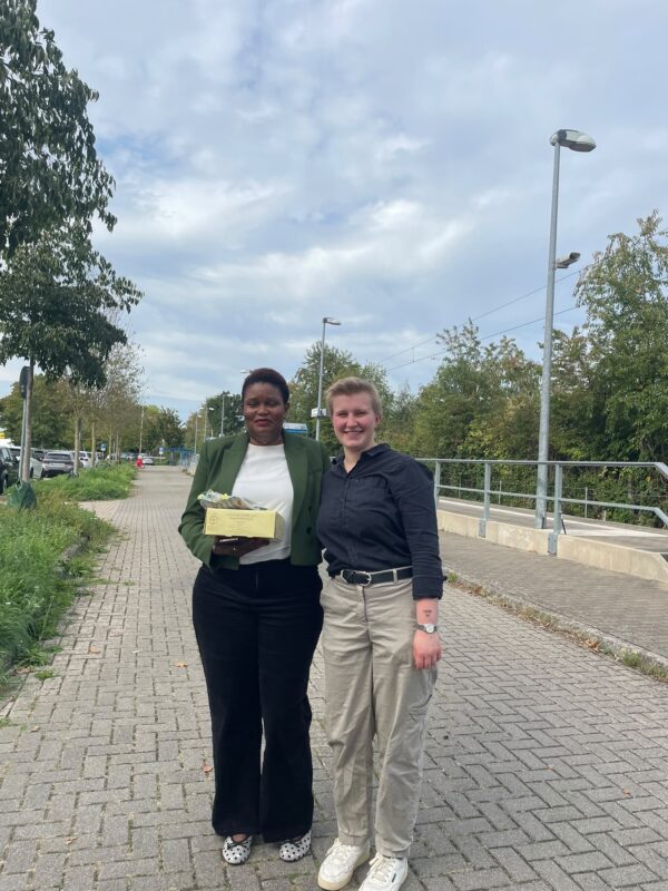 Zwei Frauen stehen lächelnd auf einem gepflasterten Weg im Freien. Links hält eine schwarze Frau in einem grünen Blazer und einem weißen Oberteil eine kleine Schachtel in der Hand. Neben ihr steht eine Person mit kurzen blonden Haaren, die ein dunkles Hemd und eine helle Hose trägt.