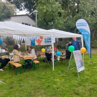 Ein Informationsstand von Plan International in einem Park, an dem Menschen an einem Tisch sitzen und ein Freiwilliger daneben steht. Auf dem Rasen stehen eine blaue Fahne und ein Schild, das für die Organisation wirbt.